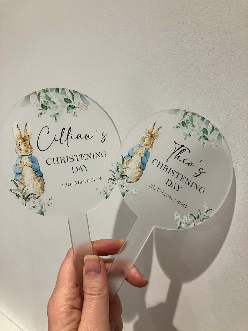 May include: Two round, clear acrylic signs with white floral designs and a watercolor illustration of Peter Rabbit. The signs have the text "Cillian's Christening Day 16th March 2024" and "Theo's Christening Day 7th February 2024".