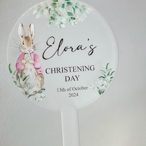May include: A white round sign with a green floral design and a cartoon rabbit. The sign says "Elora's Christening Day 13th of October 2024".