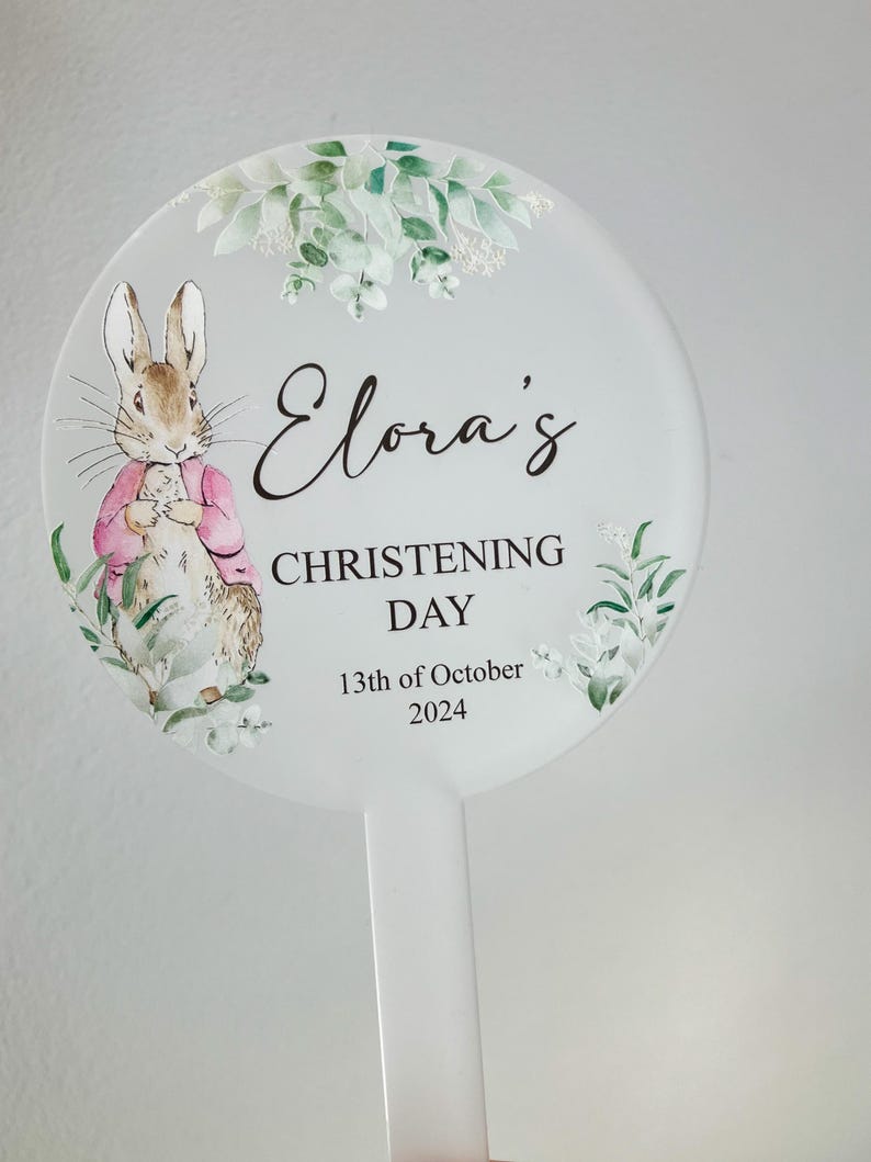 May include: A white round sign with a green floral design and a cartoon rabbit. The sign says "Elora's Christening Day 13th of October 2024".