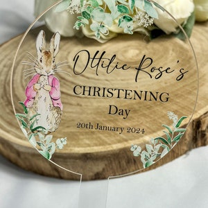 10 centimeter round clear acrylic cake topper. Decorated with images of Peter Rabbit in a pink jacket and eucalyptus foliage. The name Ottilie is printed in cursive font and the words christening day and a date are printed in capitals.
