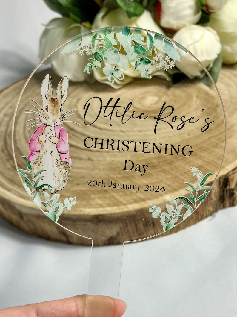 10 centimeter round clear acrylic cake topper. Decorated with images of Peter Rabbit in a pink jacket and eucalyptus foliage. The name Ottilie is printed in cursive font and the words christening day and a date are printed in capitals.