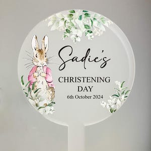May include: A white round sign with a green leafy border and a watercolor illustration of Peter Rabbit. The sign says "Sadie's Christening Day 6th October 2024".