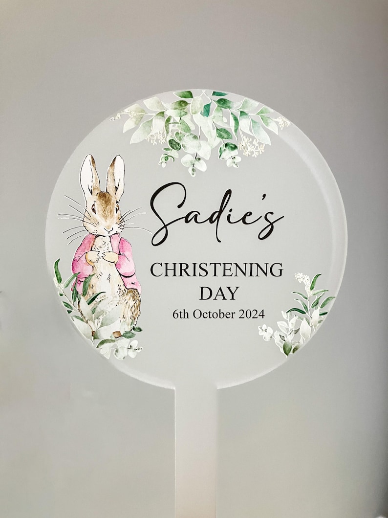 May include: A white round sign with a green leafy border and a watercolor illustration of Peter Rabbit. The sign says "Sadie's Christening Day 6th October 2024".