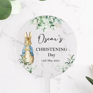 10 centimeter round clear acrylic cake topper. Decorated with images of Peter Rabbit in a blue jacket and eucalyptus foliage. The name Oscar is printed in cursive font and the words christening day and a date are printed in capitals.
