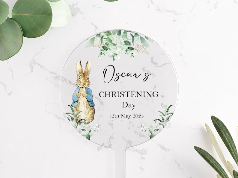 10 centimeter round clear acrylic cake topper. Decorated with images of Peter Rabbit in a blue jacket and eucalyptus foliage. The name Oscar is printed in cursive font and the words christening day and a date are printed in capitals.