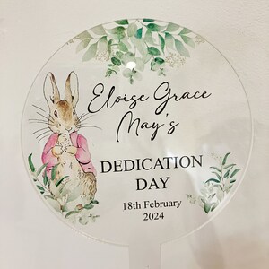 May include: A round, white, floral-patterned sign with a Peter Rabbit illustration. The sign reads "Eloise Grace May's Dedication Day 18th February 2024".