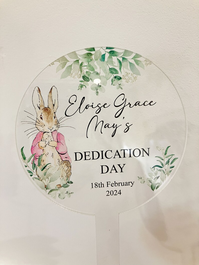 May include: A round, white, floral-patterned sign with a Peter Rabbit illustration. The sign reads "Eloise Grace May's Dedication Day 18th February 2024".