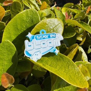 May include: A light blue plastic charm with the text "STAY IN BLOOM" in white lettering. The charm features a stylized flower design.
