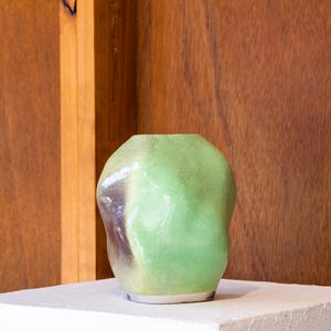 May include: A small, rounded ceramic vase with an irregular, organic shape. The surface features a mottled glaze in shades of light green, with subtle hints of dark purple and grey near the base. The opening at the top is circular and smooth.