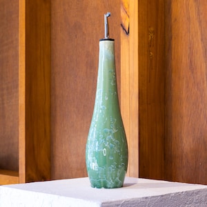 May include: A tall, slender ceramic oil cruet with a tapered neck and a wide base. The surface features a glossy, mottled sage green glaze with white crystalline patterns throughout. A silver-coloured metal pour spout is inserted into the top opening.