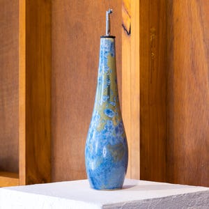 May include: A tall, slender ceramic oil bottle featuring a mottled glaze in shades of blue and tan. The bottle has a tapered neck and a wide, rounded base, topped with a silver-coloured metal pour spout with a black rubber seal at the opening.