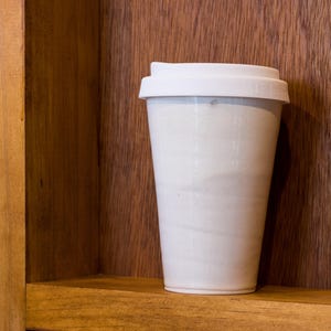 May include: A white ceramic takeaway cup with a lid on a wooden shelf.