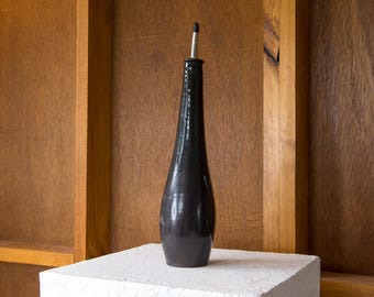May include: A tall, black, ceramic bottle with a long, slender neck and a silver and black stopper. The bottle is displayed on a white, textured surface against a wooden background.