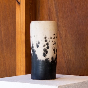 May include: A cylindrical ceramic vase featuring a two-tone colour scheme. The top portion is an off-white cream colour, while the bottom half is solid black. The transition between the two colours displays a speckled, organic pattern of black spots.