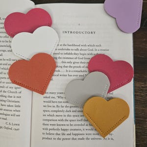 May include: Heart-shaped leather bookmarks in various colors, including pink, white, orange, gray, yellow, and purple, are placed on the pages of a book. The bookmarks have a stitched border and are used to mark pages.