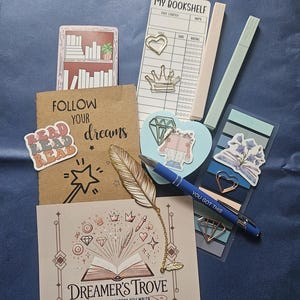 Puede incluir: Una colección de artículos de papelería, que incluye un marcapáginas, pegatinas, un bolígrafo y un bloc de notas. Los artículos presentan ilustraciones de libros, corazones y una pluma. El bloc de notas dice "MY BOOKSHELF". El bolígrafo tiene el texto "YOU GOT THIS".