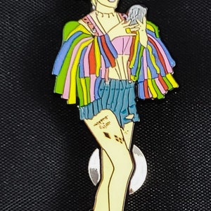 May include: A colorful enamel pin featuring a cartoon character with blonde hair, a rainbow striped jacket, blue denim shorts, and white and pink boots. The character is holding a small, round object in their hand. The pin has a silver backing.