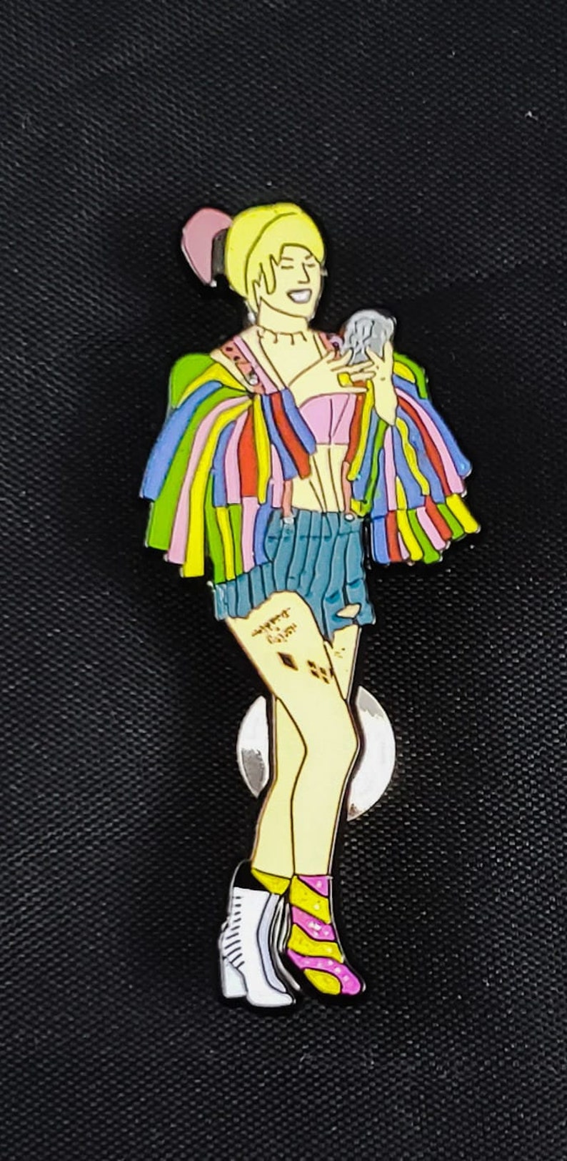 May include: A colorful enamel pin featuring a cartoon character with blonde hair, a rainbow striped jacket, blue denim shorts, and white and pink boots. The character is holding a small, round object in their hand. The pin has a silver backing.