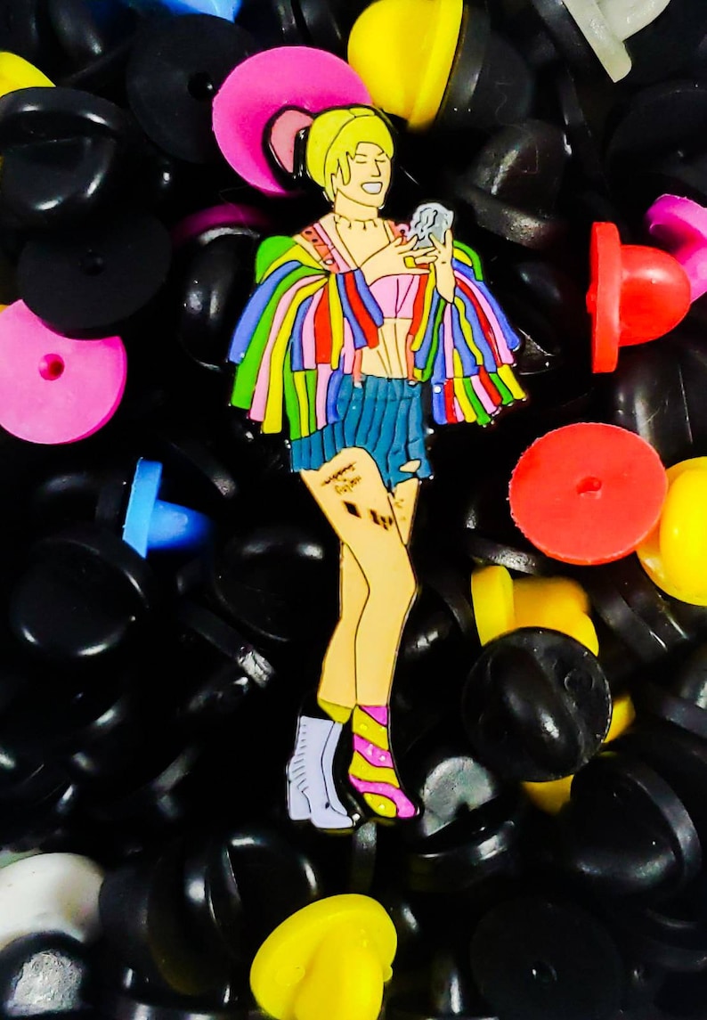 May include: A colorful enamel pin featuring a woman with blonde hair wearing a rainbow striped jacket, a pink top, blue denim shorts, and white and pink striped socks. She is holding a microphone in her right hand. The pin is surrounded by black, yellow, red, and blue plastic push pins.