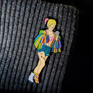 May include: A colorful enamel pin featuring a cartoon character with blonde hair, wearing a rainbow-colored jacket, denim shorts, and white boots. The character is holding a small, white object in their right hand.