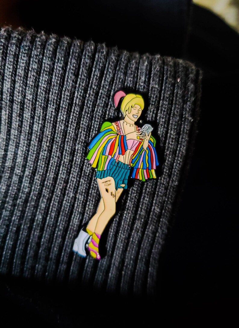 May include: A colorful enamel pin featuring a cartoon character with blonde hair, wearing a rainbow-colored jacket, denim shorts, and white boots. The character is holding a small, white object in their right hand.