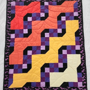 Könnte beinhalten: Ein farbenfroher Quilt mit einem geometrischen Muster in Orange-, Gelb-, Lila- und Schwarztönen. Der Quilt ist mit einem schwarzen Stoff mit einem sich wiederholenden Muster aus Fledermäusen eingefasst.