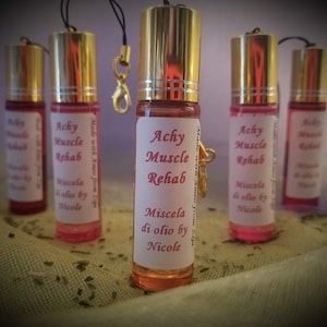 Puede incluir: Cinco pequeños frascos de vidrio con tapas doradas y etiquetas rosas. Las etiquetas dicen "Achy Muscle Rehab" y "Miscela di olio by Nicole".