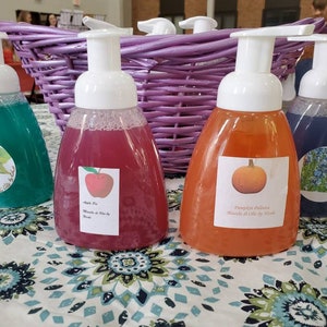 Pode incluir: Quatro garrafas de sabonete de mãos espumante com diferentes aromas e rótulos. Os aromas são especiarias de abóbora, bordo e noz-pecã, hortelã-pimenta e lavanda. As garrafas são transparentes e têm bombas brancas. As garrafas estão sobre uma mesa com um padrão floral.