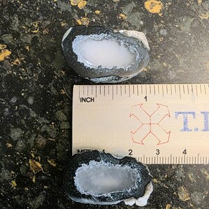 May include: Two halves of a geode, each with a white crystal center and a dark gray outer edge. A ruler shows the geodes are approximately 1.5 inches in length. The background is a dark, speckled surface.