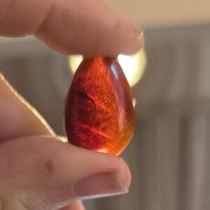 May include: A teardrop-shaped, translucent pendant with a gradient of red and orange hues. The pendant is held between two fingers, showcasing its polished surface and internal marbling effect. The background is blurred.