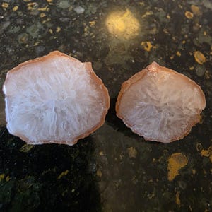 May include: Two agate slices with a light pink and white color scheme. The edges are a darker shade of pink, and the interior has a crystalline appearance. The slices are irregularly shaped and appear to be polished.