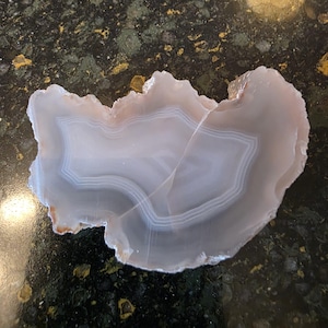 May include: A polished agate slice with a natural, irregular edge. The stone features concentric bands of white, gray, and pale blue, creating a layered effect. The agate has a translucent quality, revealing its internal structure.