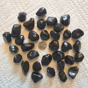 May include: A collection of polished black obsidian stones of various shapes and sizes. A US dime is included for size reference. The stones have a glossy, reflective surface and are arranged on a white, textured surface.