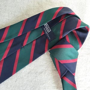 May include: A striped necktie in navy, green, and red. The tie features diagonal stripes and a label that reads "POLO by Ralph Lauren". The tie is knotted and lies on a textured surface.