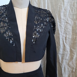May include: Black cropped jacket with long sleeves and a sequined design on the shoulders and upper chest. The jacket is displayed on a mannequin. The jacket has a V-neck and is open in the front.