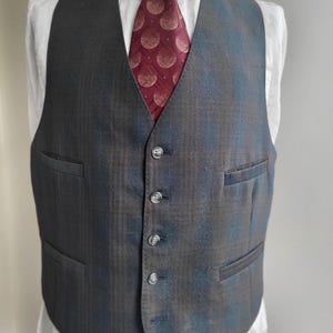 May include: A grey waistcoat with a subtle plaid pattern, featuring a V-neck and four buttons. A burgundy tie with a repeating circular design is visible, paired with a white collared shirt. The waistcoat has two front pockets.