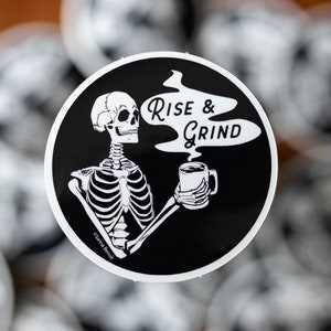 Sticker of a skeleton holding a coffee mug and in the steam coming off the coffee is text reading Rise & Grind