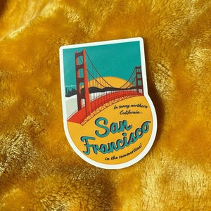 Sticker with a teal sky and yellow sun with the red Golden Gate Bridge. In teal script lettering it reads, San Francisco, and around it reads, In sunny northern California... in the summertime! Sticker rests on a yellow blanket.