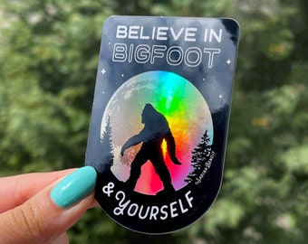 May include: A black and white sticker with a holographic moon and a silhouette of Bigfoot. The text on the sticker reads "Believe in Bigfoot & Yourself".