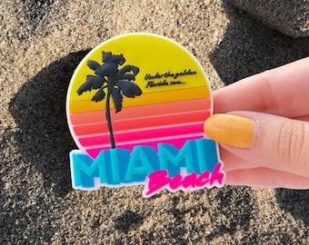 May include: A yellow, pink, and blue Miami Beach magnet with a palm tree silhouette and the text "Under the golden Florida sun..." and "Miami Beach".