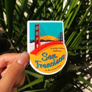 Sticker with a teal sky and yellow sun with the red Golden Gate Bridge. In teal script lettering it reads, San Francisco, and around it reads, In sunny northern California... in the summertime! Fingers hold the sticker in front of a palm bush.