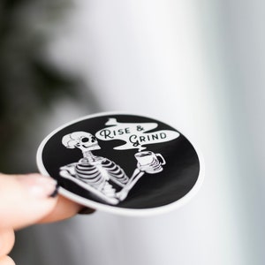 Sticker of a skeleton holding a coffee mug and in the steam coming off the coffee is text reading Rise & Grind