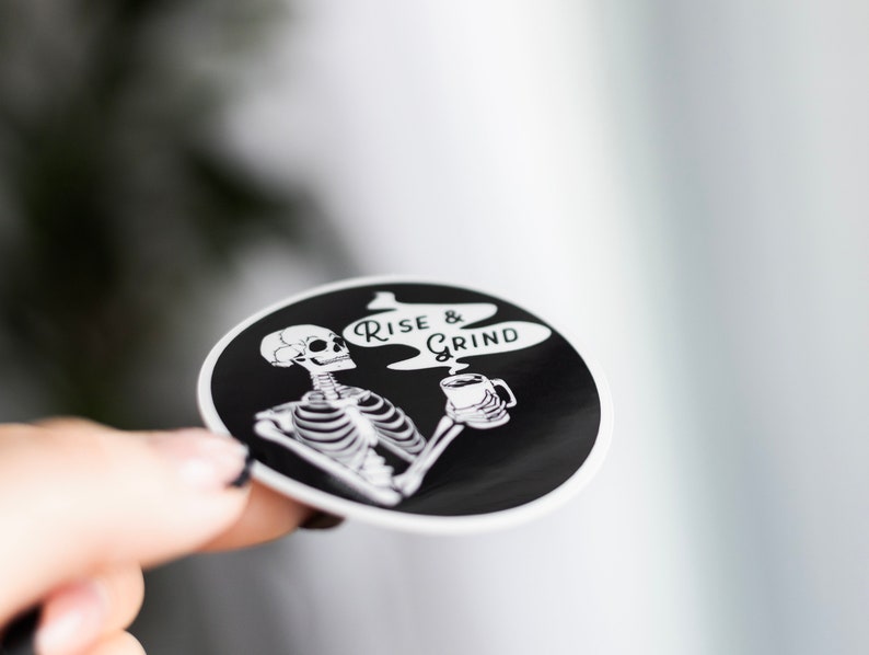 Sticker of a skeleton holding a coffee mug and in the steam coming off the coffee is text reading Rise & Grind