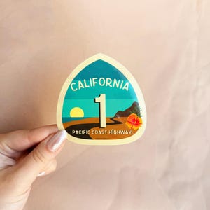 May include: A yellow and blue sticker with the text "California 1 Pacific Coast Highway". The sticker features a sunset scene with a palm tree and a road.