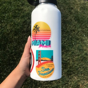 Sticker with a teal sky and yellow sun with the red Golden Gate Bridge. In teal script lettering it reads, San Francisco, and around it reads, In sunny northern California... in the summertime! Sticker is on white water bottle held above grass.