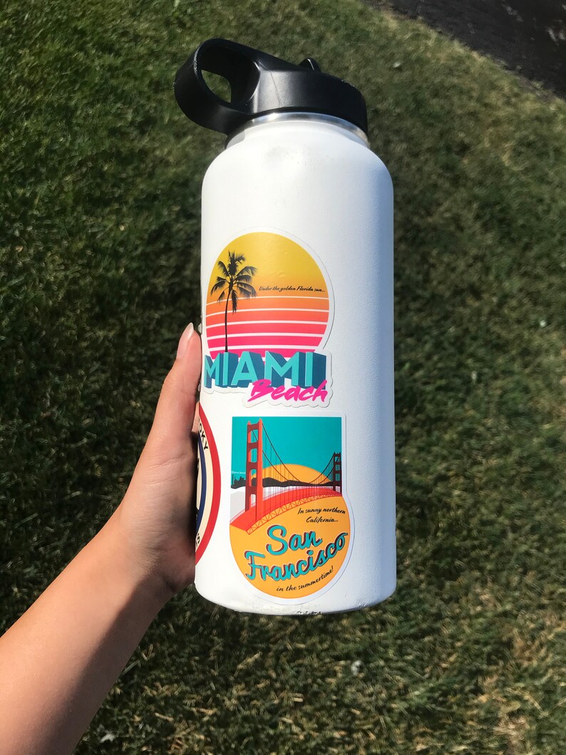 Sticker with a teal sky and yellow sun with the red Golden Gate Bridge. In teal script lettering it reads, San Francisco, and around it reads, In sunny northern California... in the summertime! Sticker is on white water bottle held above grass.