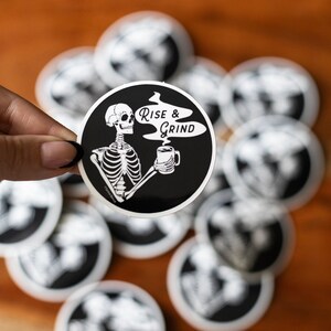 Sticker of a skeleton holding a coffee mug and in the steam coming off the coffee is text reading Rise & Grind