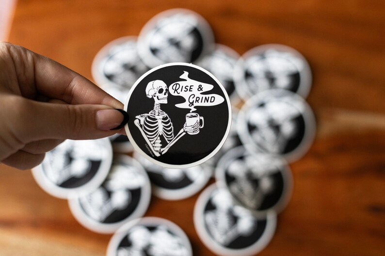 Sticker of a skeleton holding a coffee mug and in the steam coming off the coffee is text reading Rise & Grind