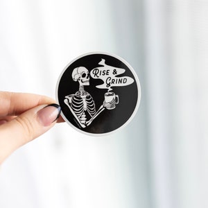 Sticker of a skeleton holding a coffee mug and in the steam coming off the coffee is text reading Rise & Grind