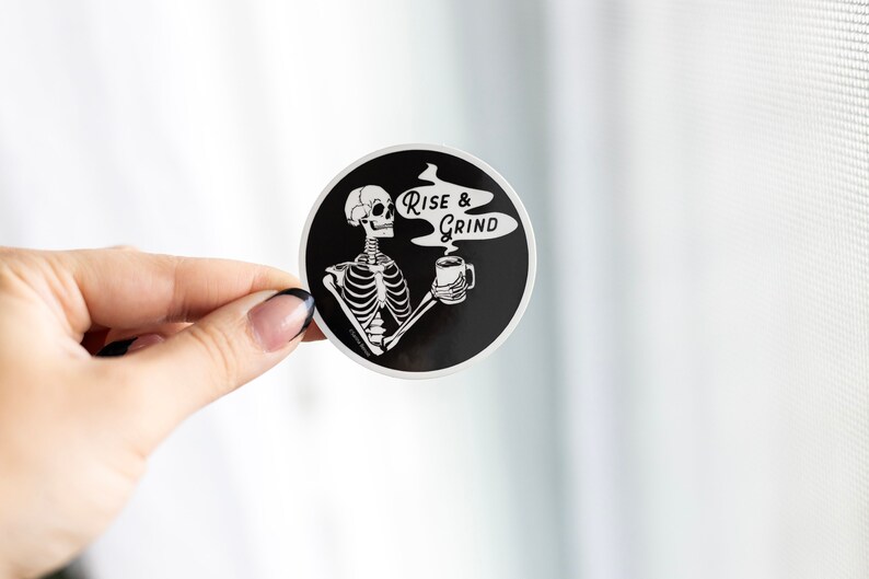 Sticker of a skeleton holding a coffee mug and in the steam coming off the coffee is text reading Rise & Grind